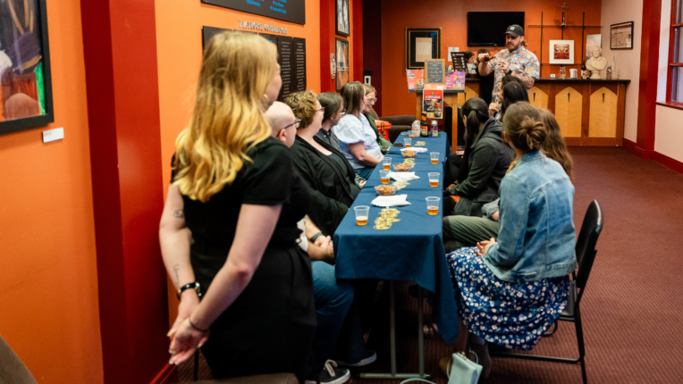 Attendees at Shakespeare Uncorked 2023 sit at a long table with small beer samples and snacks, listening to a beer expert discuss Devil’s Backbone brews at the American Shakespeare Center.