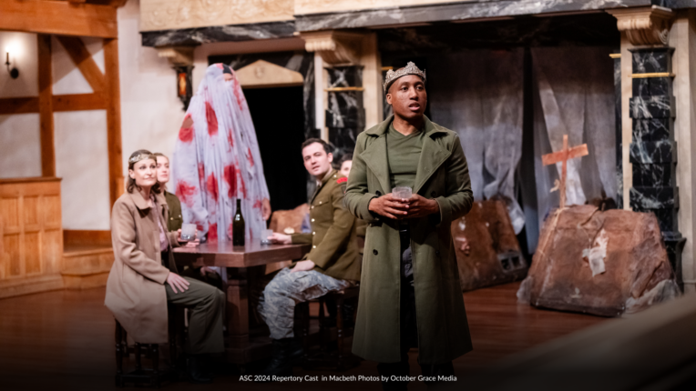 K.P. Powell as Macbeth stands in front of a banquet table with the 2024 ASC Repertory Cast, while Kenn Hopkins Jr. as Banquo’s ghost looms in the background. The scene unfolds on the Blackfriars Playhouse stage, emphasizing the modern military setting of the production.