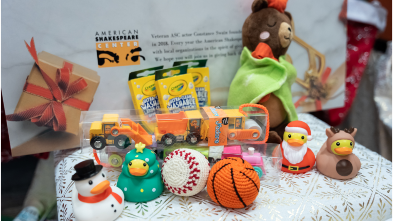 A variety of holiday gifts, including Crayola markers, toy construction vehicles, festive rubber ducks (decorated as Santa, a reindeer, and a Christmas tree), and crocheted balls resembling a baseball and basketball, displayed on a patterned tablecloth for Project Goodfellow at the American Shakespeaere Center.