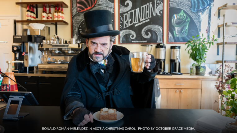 Ronald Román-Meléndez as Ebenezer Scrooge in A Christmas Carol at the American Shakespeare Center, holding a glass of Hot Apple Wassail and a plate of Apple Pie Bostock at Reunion Bakery in Staunton, Virginia. The cozy bakery background features coffee machines, plants, and a chalkboard menu.