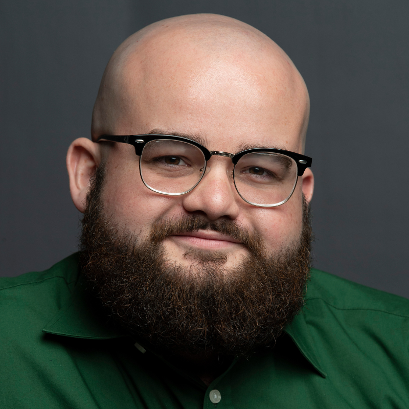 Braden Miller Portrait of Braden Miller, Props Artisan at the American Shakespeare Center, wearing glasses and a green shirt against a dark background.