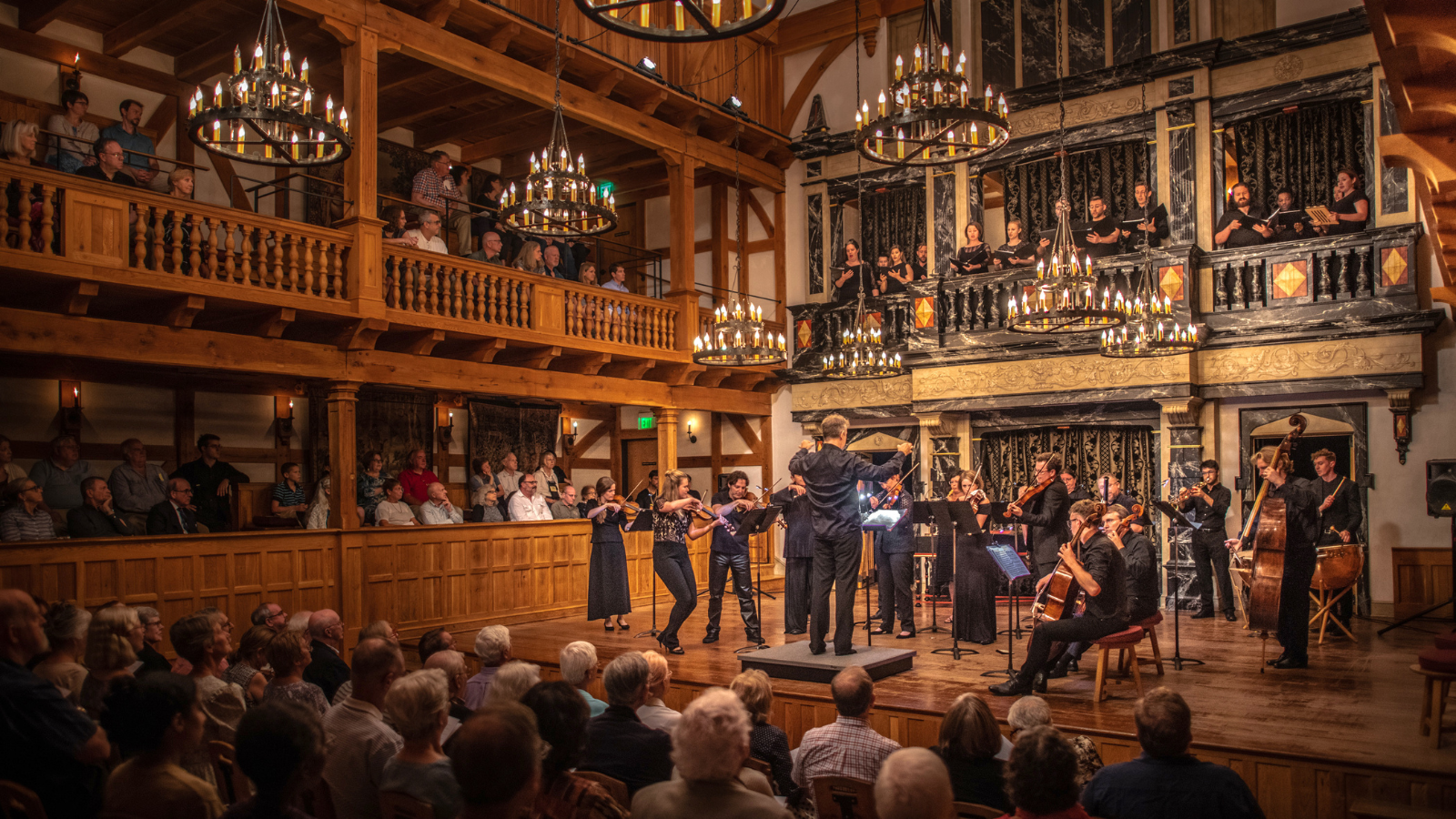 A live performance by Staunton Music Festival inside the Blackfriars Playhouse in Staunton, Virginia. Musicians and vocalists fill the stage beneath glowing chandeliers, surrounded by a full audience in the historic, wood-paneled theatre.
